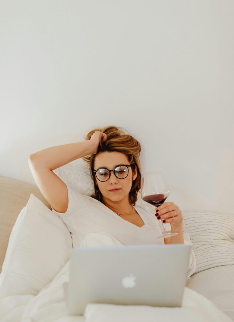 Woman lying on bed with laptop and glass of wine, looking overwhelmed and reflective, representing feeling stuck in life despite being capable and ambitious.