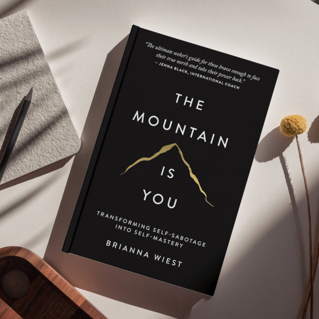 Flat lay of the book The Mountain Is You: Transforming Self-Sabotage Into Self-Mastery by Brianna Wiest, featuring a black minimalist cover with gold mountain line art, placed on a neutral surface alongside a notebook, pencil, and dried floral stem.