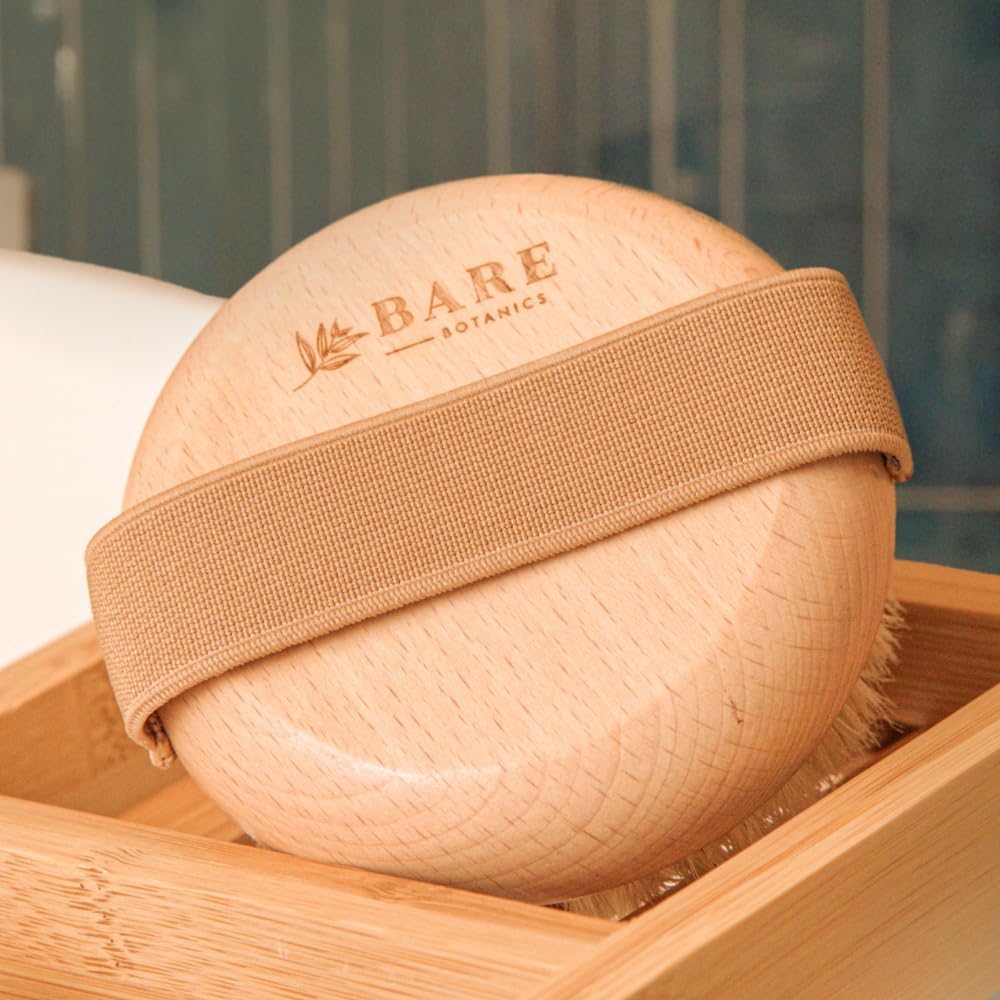 Wooden body dry brush with natural bristles and a cotton hand strap, engraved with Bare Botanics logo, resting in a bamboo tray for exfoliation and lymphatic massage.
