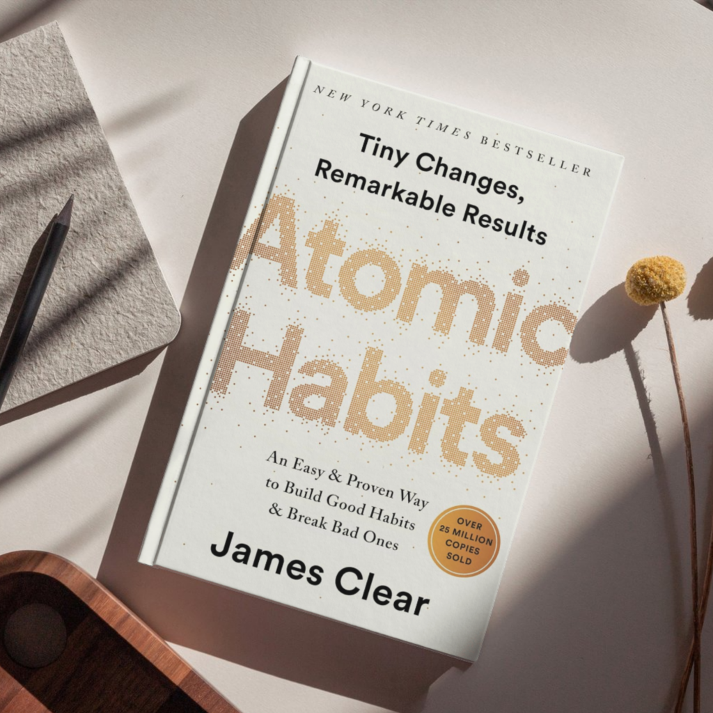 Flat lay of the book Atomic Habits: Tiny Changes, Remarkable Results by James Clear, featuring a white minimalist cover with gold dotted typography, placed on a neutral surface alongside a notebook, pencil, wooden tray, and dried floral stem.