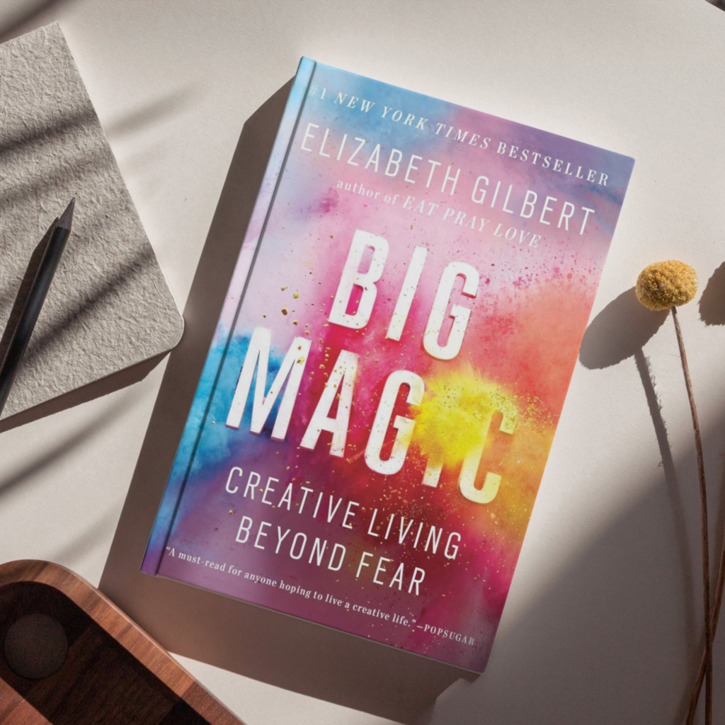 Flat lay of the book Big Magic: Creative Living Beyond Fear by Elizabeth Gilbert, featuring a colorful watercolor-style cover, placed on a neutral surface alongside a notebook, pencil, wooden tray, and dried floral stem.