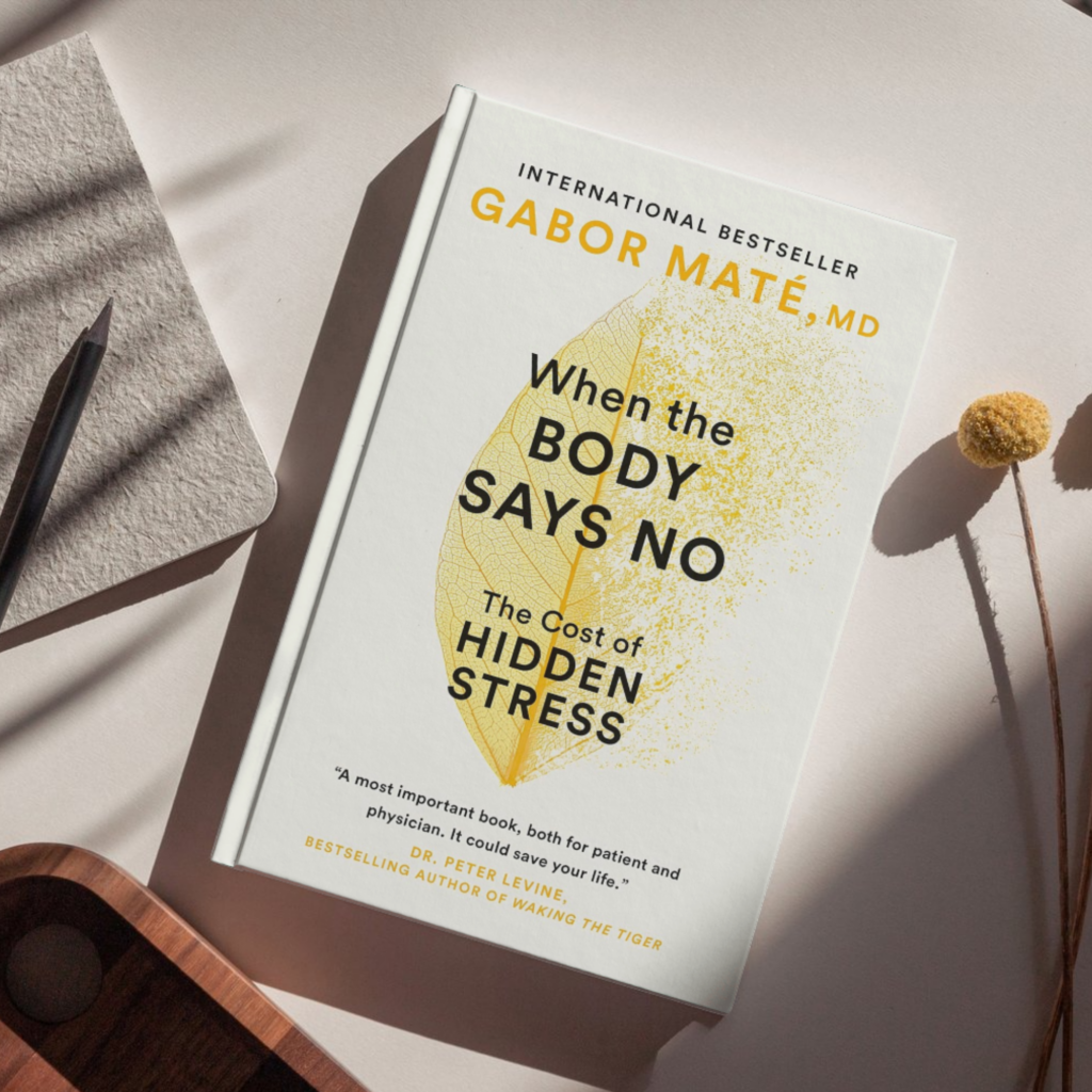 lat lay of the book When the Body Says No: The Cost of Hidden Stress by Gabor Maté, featuring a minimalist white cover with a yellow leaf illustration, placed on a neutral surface alongside a notebook, pencil, wooden tray, and dried floral stem.
