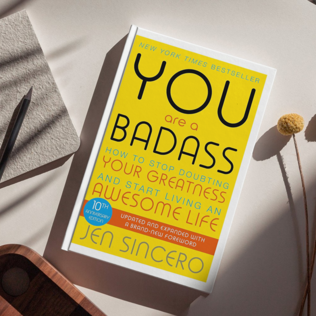 Flat lay of the book You Are a Badass: How to Stop Doubting Your Greatness and Start Living an Awesome Life by Jen Sincero, featuring a bright yellow cover with bold typography, placed on a neutral surface alongside a notebook, pencil, wooden tray, and dried floral stem.